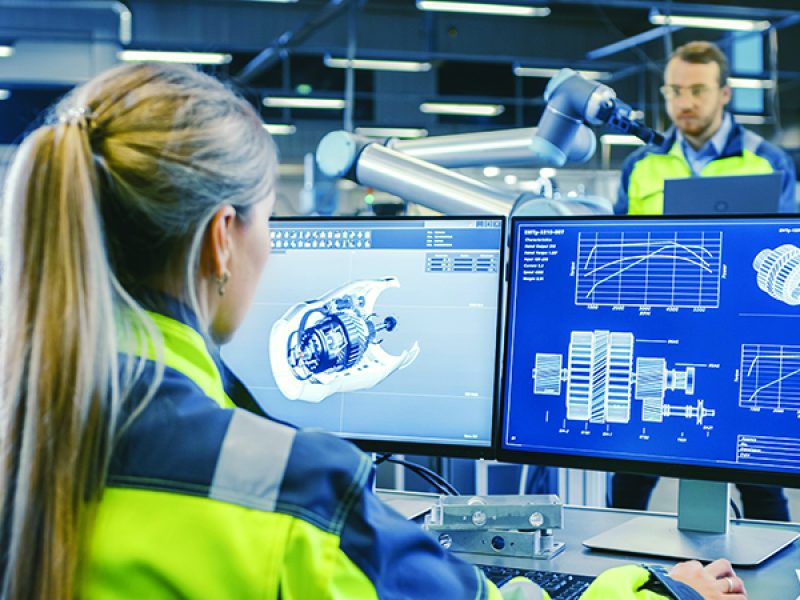 At the Factory: Female Mechanical Engineer Designs 3D Engine on Her Personal Computer. In the Background Male Automation Engineer who Uses Laptop for Programming Robotic Arm.